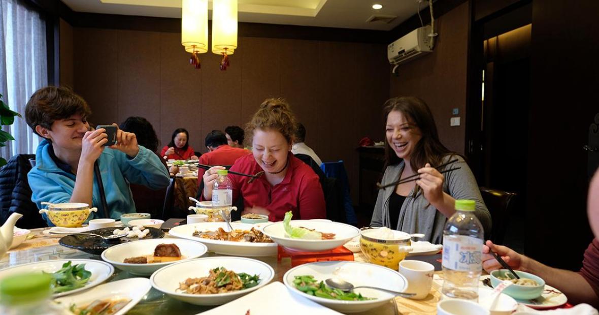 Two diners laughing while lifting eels with chopsticks. Table loaded with a variety of food.