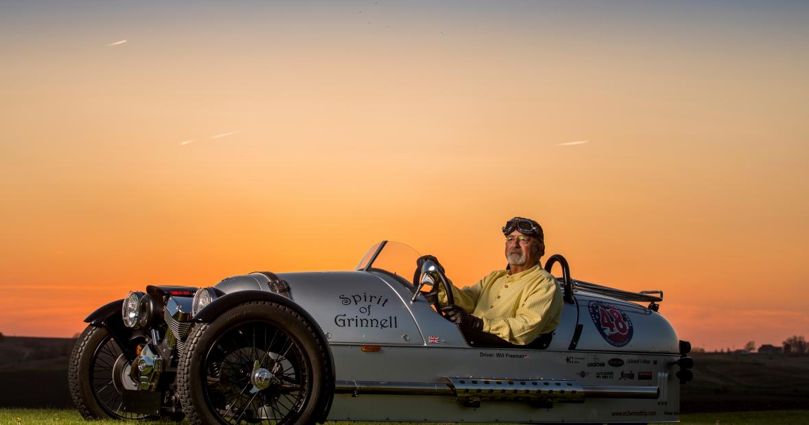 Will Freeman sits in his Morgan-Three Wheeler, named The Spirit of Grinnell, as the sun sets behind him
