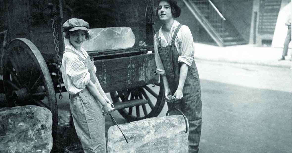 Two young women carry a large block of ice between them with large tongs