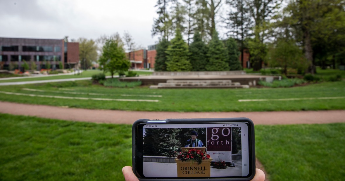 Smart phone in foreground showing virtual commencement in front of empty outdoor commencement stage in background