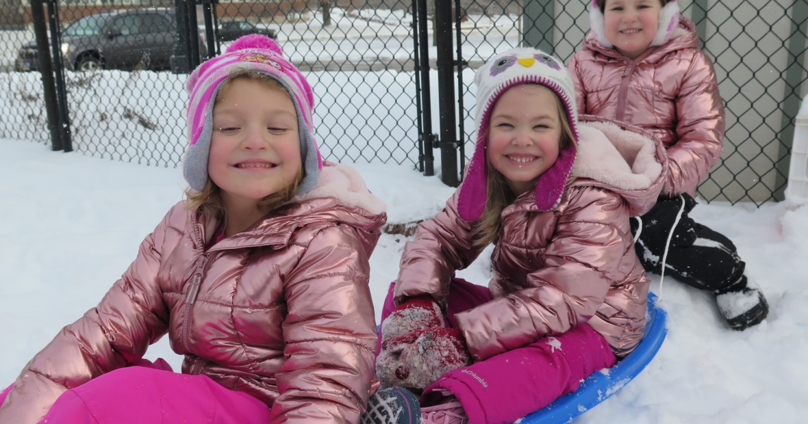 three preschool students sitting in the snow.  Two students are sitting in a blue sled.