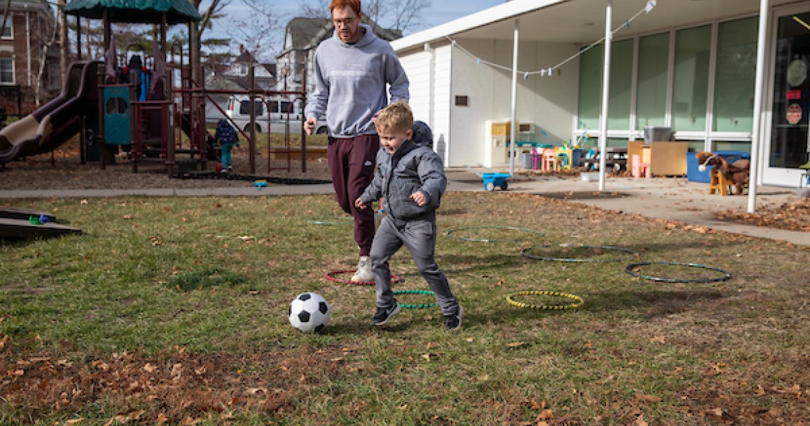 child plays soccer with caregiver at preschool playground