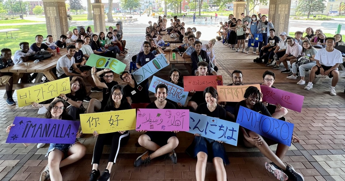 Several students sitting in a park shelter, smiling and holding signs with welcome in different languages