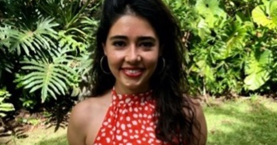 Lorena is smiling and wearing a red dress, with foliage in the background. 