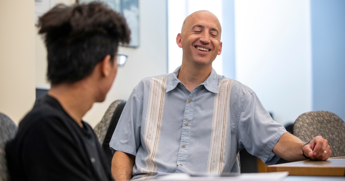 Jeff Blanchard, in a blue button-down shirt, laughs while sitting at a table with a student.