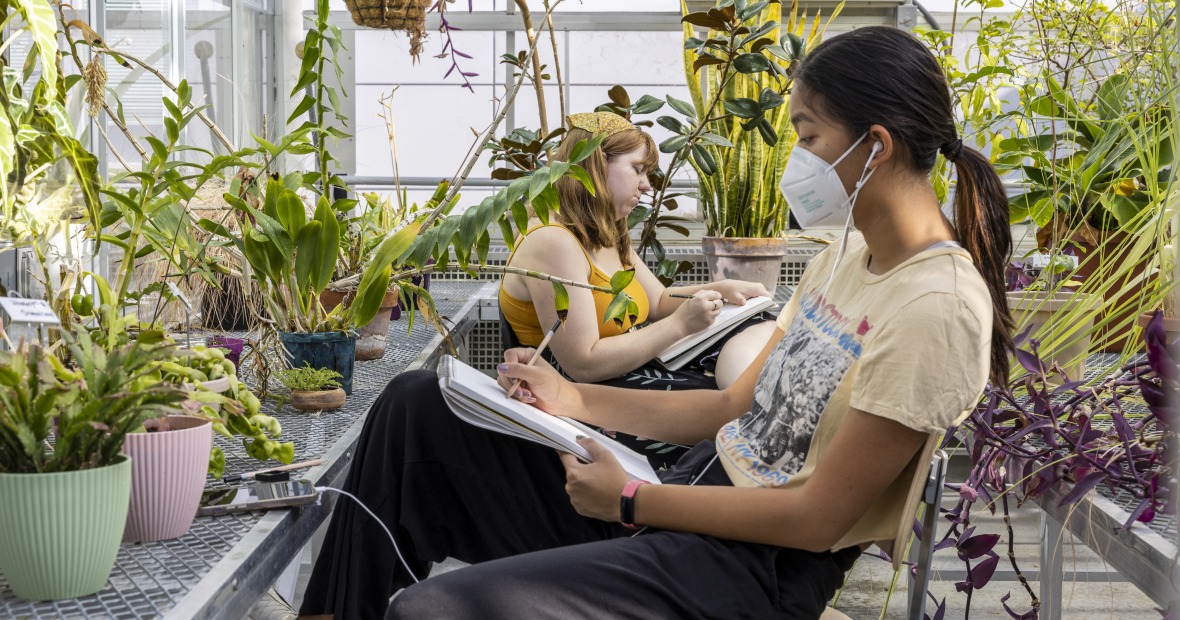 Two students sit between rows of plants in the greenhouse and sketch.