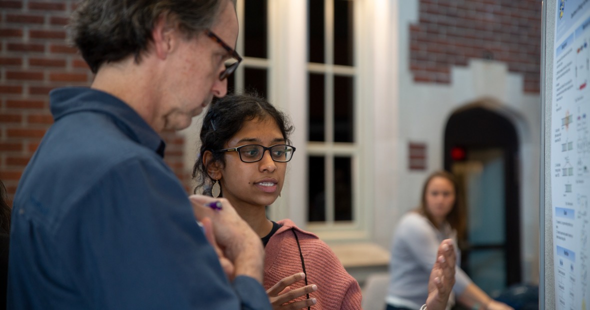 A young woman in an orange shirt explains her research poster to a man in a blue shirt, who is listening.