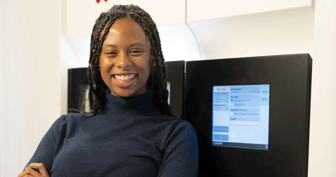 A young woman in a dark blue turtleneck and braided hair crosses her arms across her chest and smiles wildly. She stands in front of a large, cryogenic electron microscope.