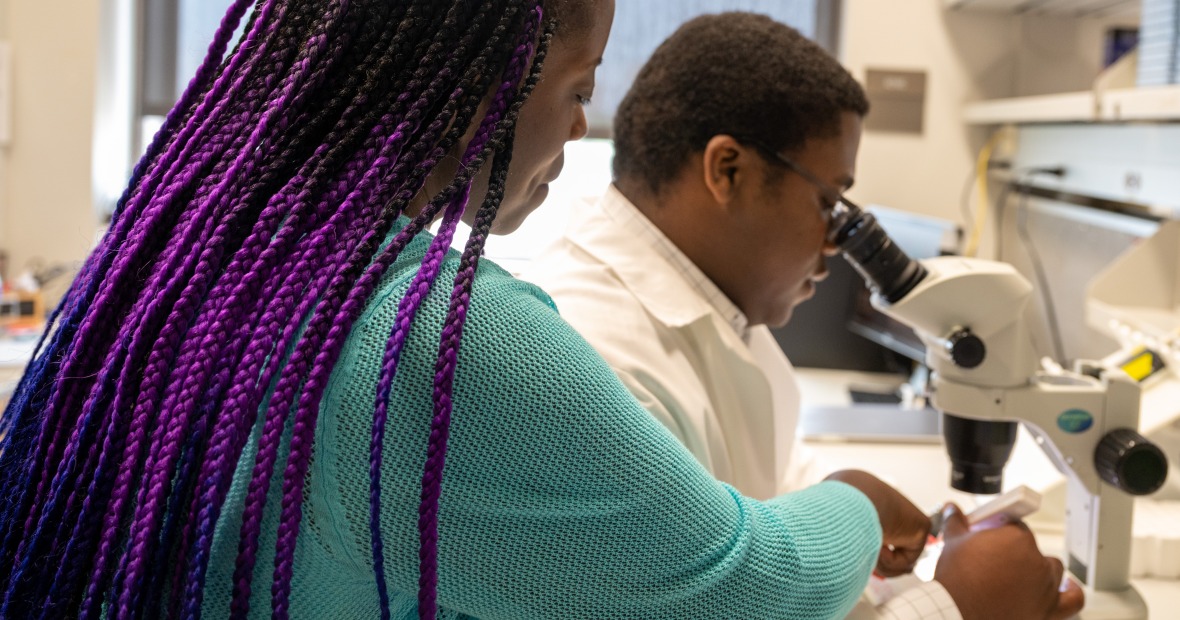 A woman with purple braids and a blue sweater looks over the shoulder of a student. She is pointing something out in the cells that the student is viewing through a microscope.