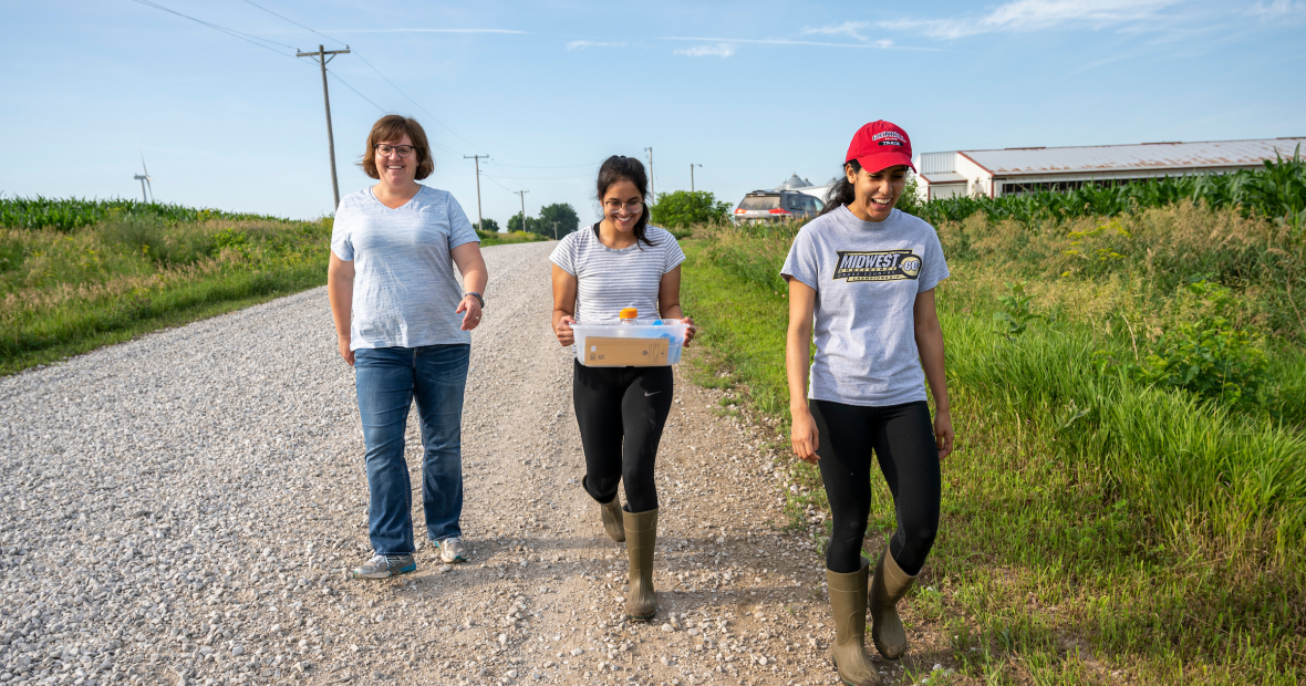 A woman and two students, also young women, walk toward the camera on a gravel road next to a field. They are conversing and smiling.