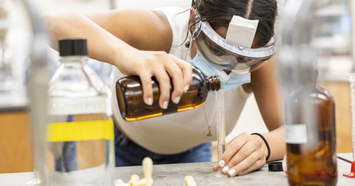 A girl with laboratory goggles leans over a bench, carefully pouring a reagent from a brown bottle into a graduated cylinder.