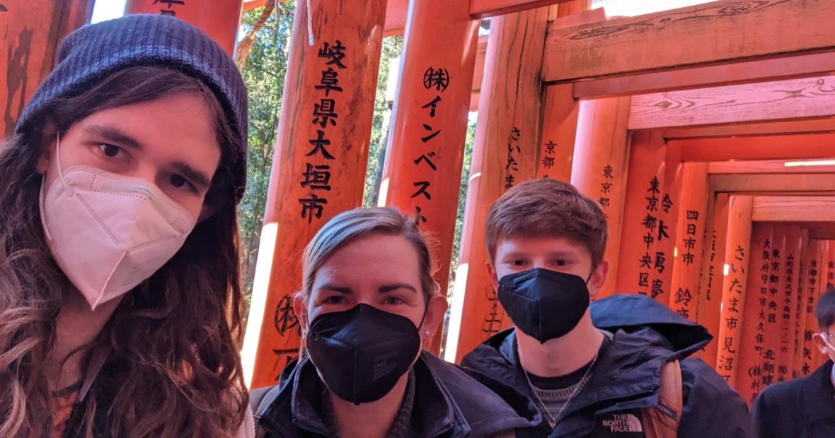 A selfie taken by three people in winter jackets and face masks. behind them are red, wooden gates inscribed with Japanese calligraphy.