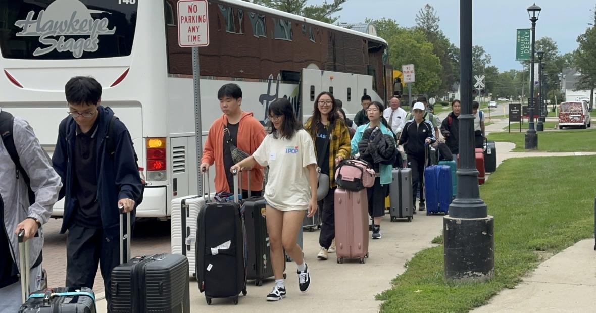 Many people walk towards the viewer, away from a large white bus, and carry luggage with them