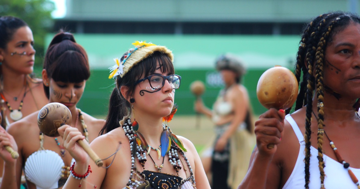 a woman with glasses and face paint  and adorned with bright feathers and beads stands in a line of women who all hold up similar instruments