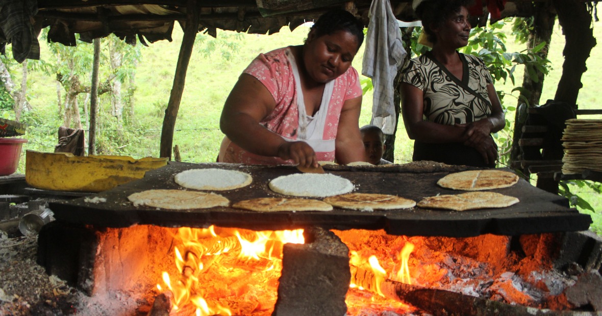 A woman standing in an open shelter uses her fingers to flip flatbread on a wide griddle stretched over a long fire
