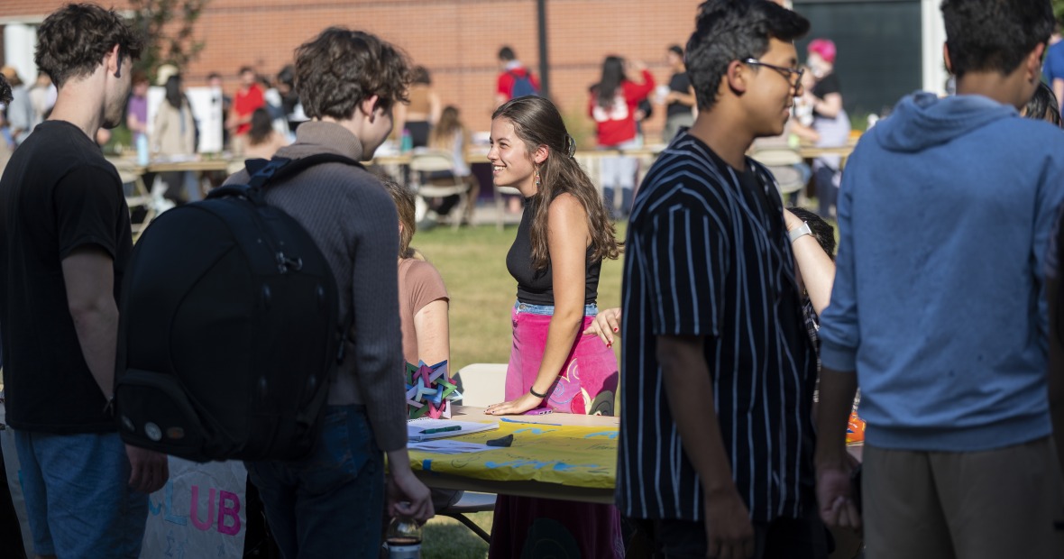 A focus on a student wearing pink velvet jeans. They talk to a student at a table outdoors.