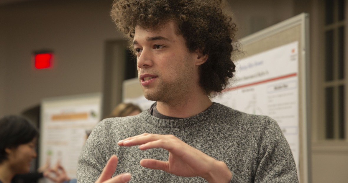 Mikey, a young man with curly brown hair, gestures while giving a presentation at a poster session.