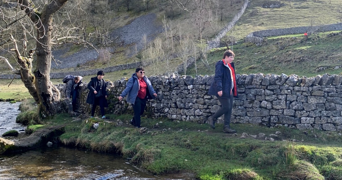 4 students, some carrying packs, walk on a grassy bank between a shallow stream and stone wall in Yorkshire Dales, England, 