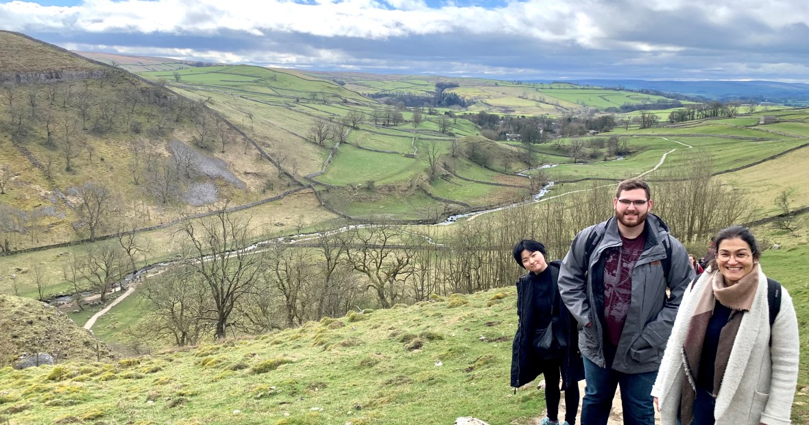 Three people, with others behind them on the path, pose with a valley and hills in the background. Faculty and students hiking to top of Malham Cove