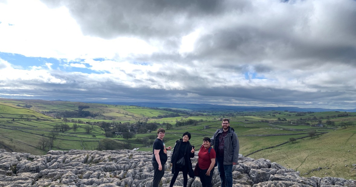 4 students standing on the rocky surface at the top of Malham Cove with rolling hills covered with fields extending in the distance behind them