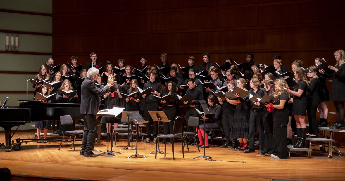 A choir sings on risers onstage, led by a male conductor