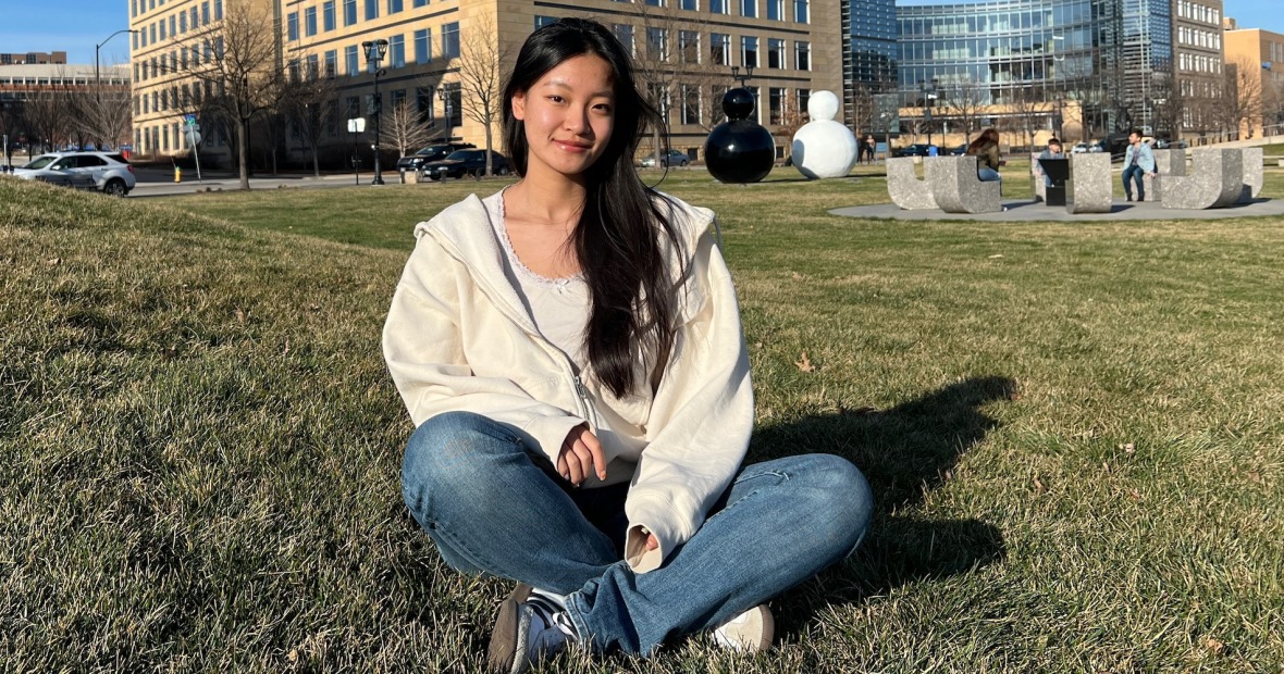 Olivia sits cross legged in a grassy area in front of a large building, smiling at the viewer