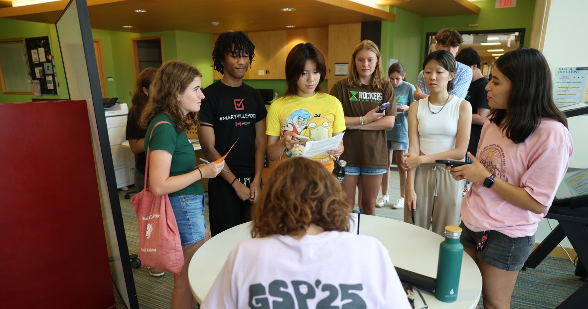 Six students stand at a low circular table where a person sits in front of them wearing a shirt that says "GSP '25"
