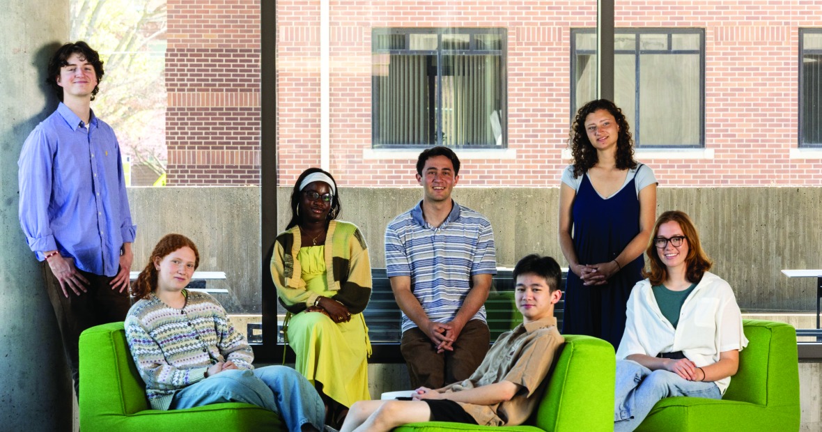 Seven class of 2025 students pose in front of a glass wall. Some stand, while others are seated, with three in low, bright green chairs.