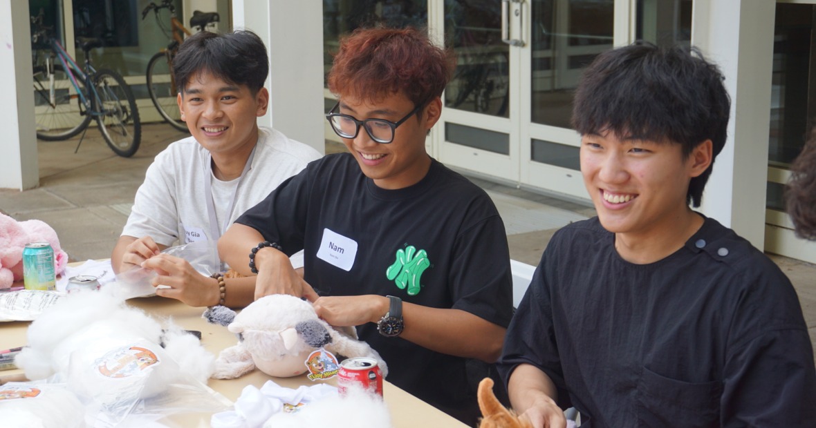 Three people sit at a table and make stuffed animals. They look off to their left and smile.