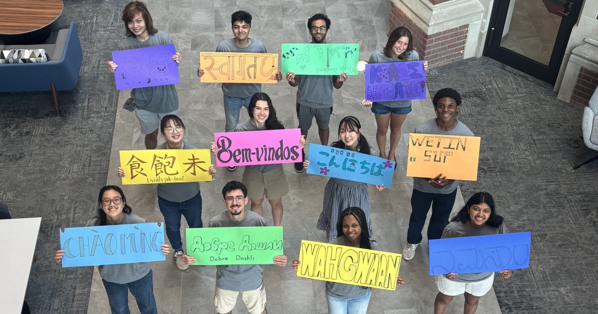 Several people hold up signs with "welcome" written in different languages and smile up at the viewer