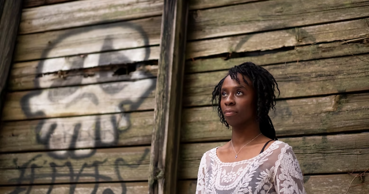 A young woman poses against a rough wooden wall painted with graffiti 