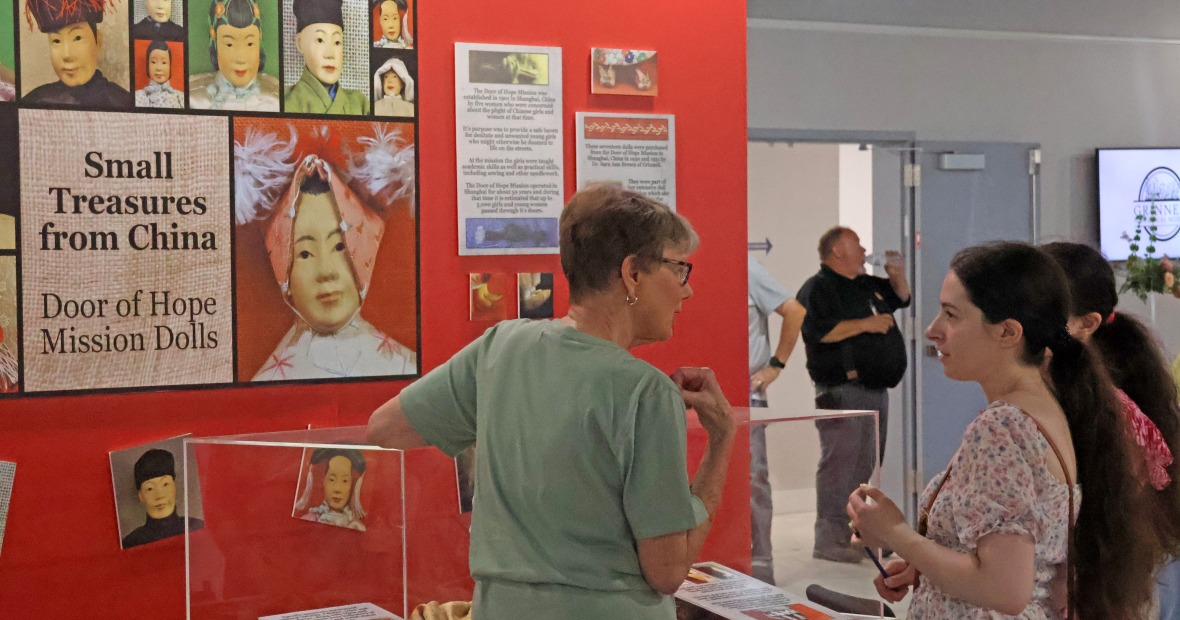 Three people stand at a glass museum case in front of a red wall with a large poster that reads 'Small Treasures from China: Door of Hope Mission Dolls'