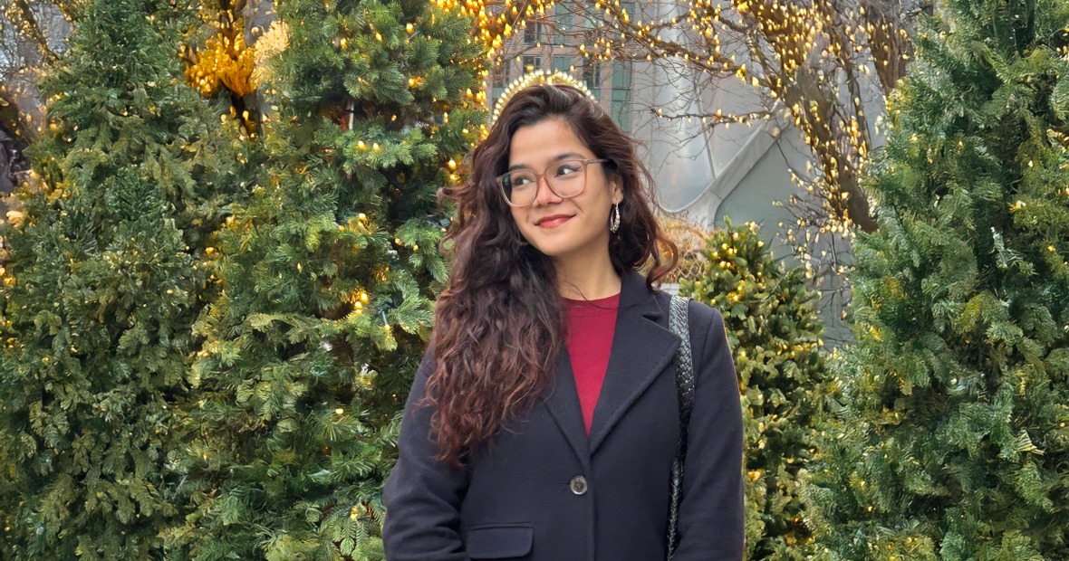 Meghna stands in front of a number of trees, some lit with white holiday lights