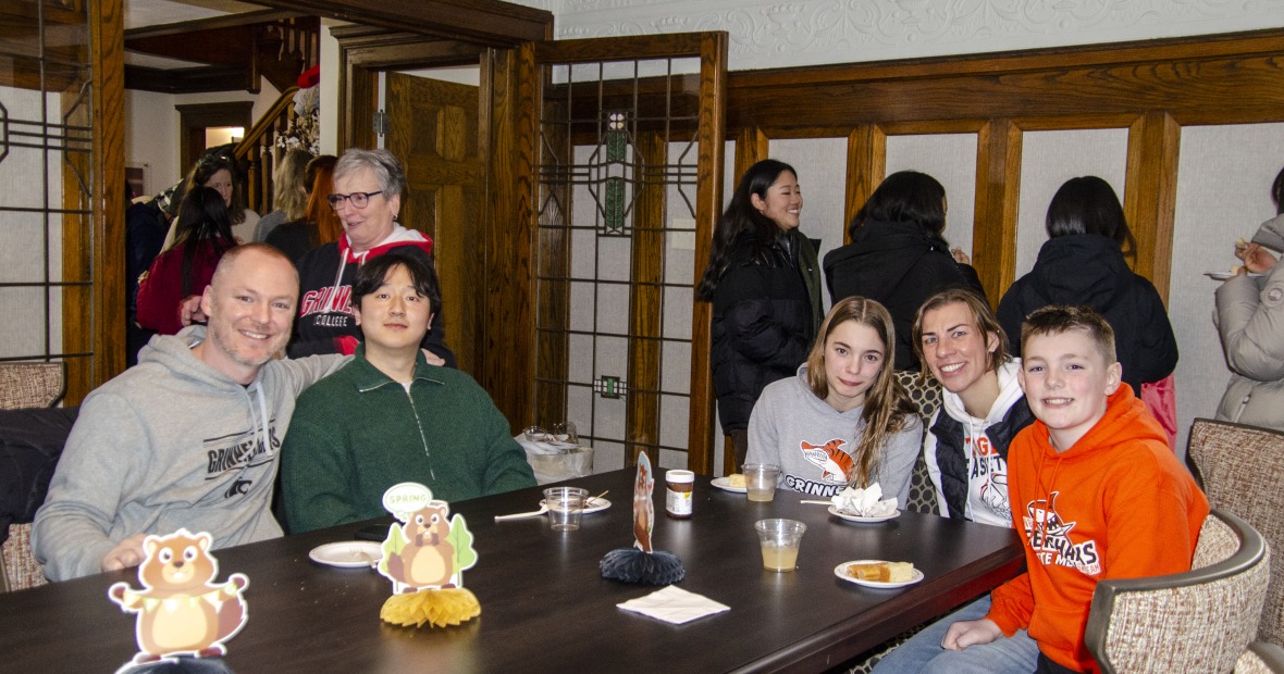 Five people sitting at a table and smiling. More people stand in the background chatting