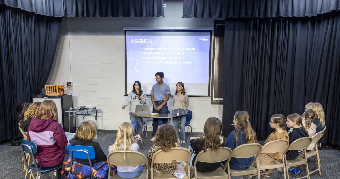 Three college students stand facing a semicircle of 15 middle school girls sitting in folding chairs