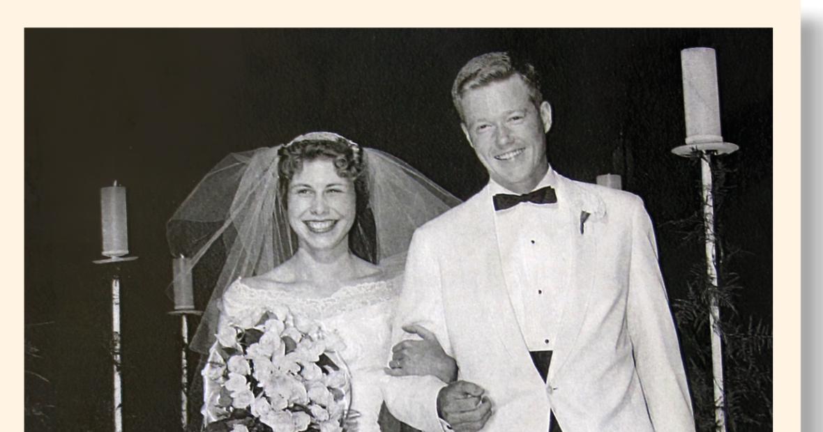 In the aisle of a church, Susan Ratcliff stands to the left in her bridal gown and a large bouquet of flowers. George Drake ihe groom stands to the right in a white jacket and black trousers.