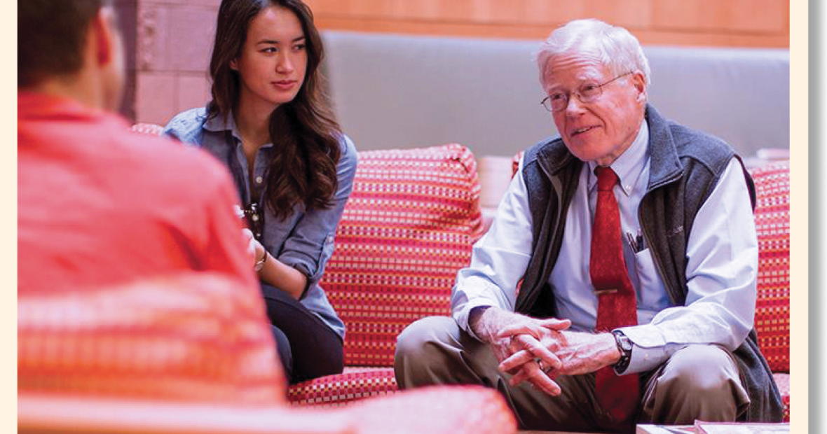 George Drake talks to a couple of students. One student sits beside him on a couch, and another across a small table.