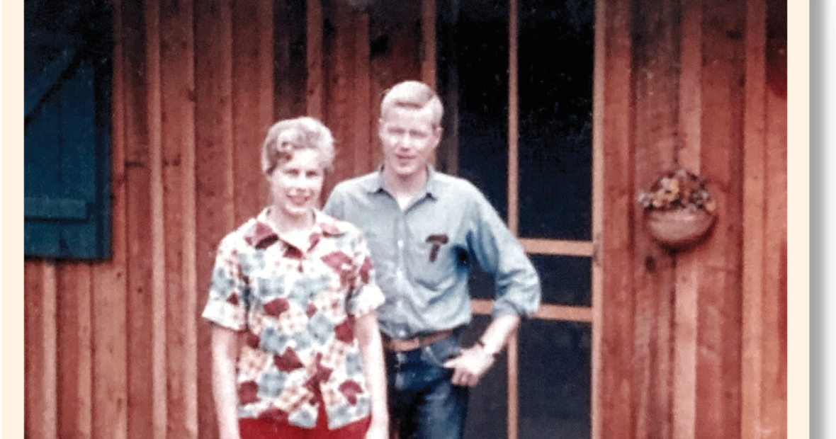 Sue and George Drake stand in front of the screen door to a cabin. The cabin has a stone stoop and black shutters, and a small basket of flowers hanging near the door.. 