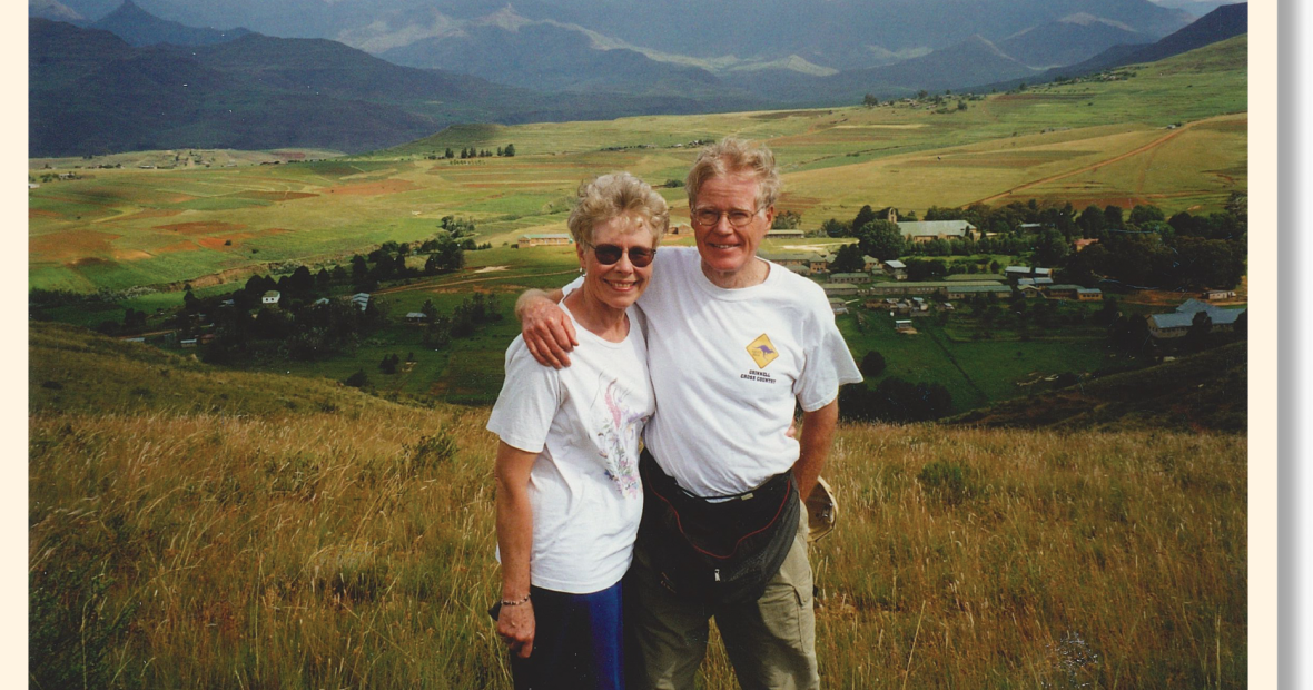 George Drake has his arm around Sue. They are both smiling in T-shirts with a broad expanse of grass and a small village visible in a shallow valley behind them.
