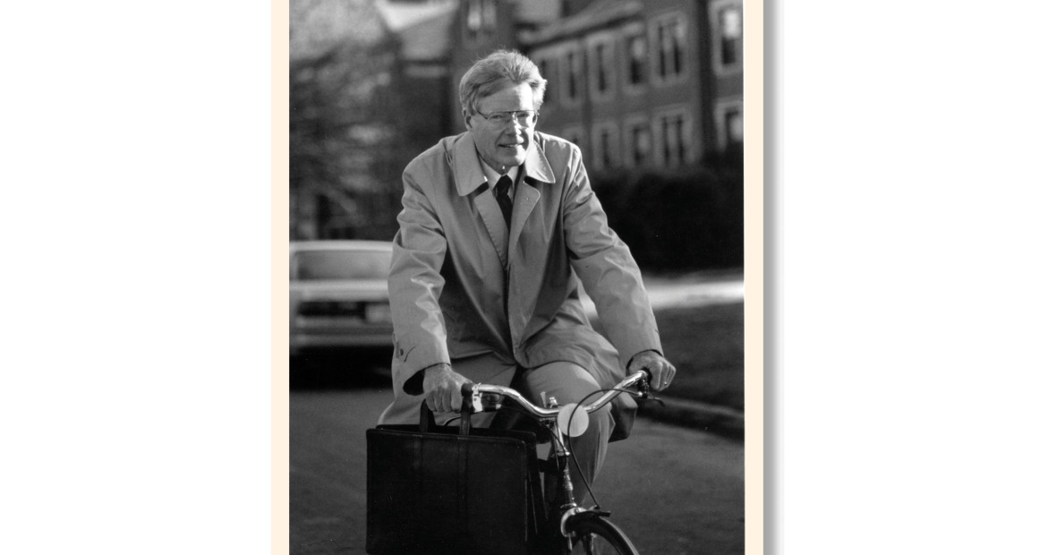 George Drake rides a bike. He is wearing a coat and clasps the handles of a leather briefcase in his right hand. North Campus is visible behind him