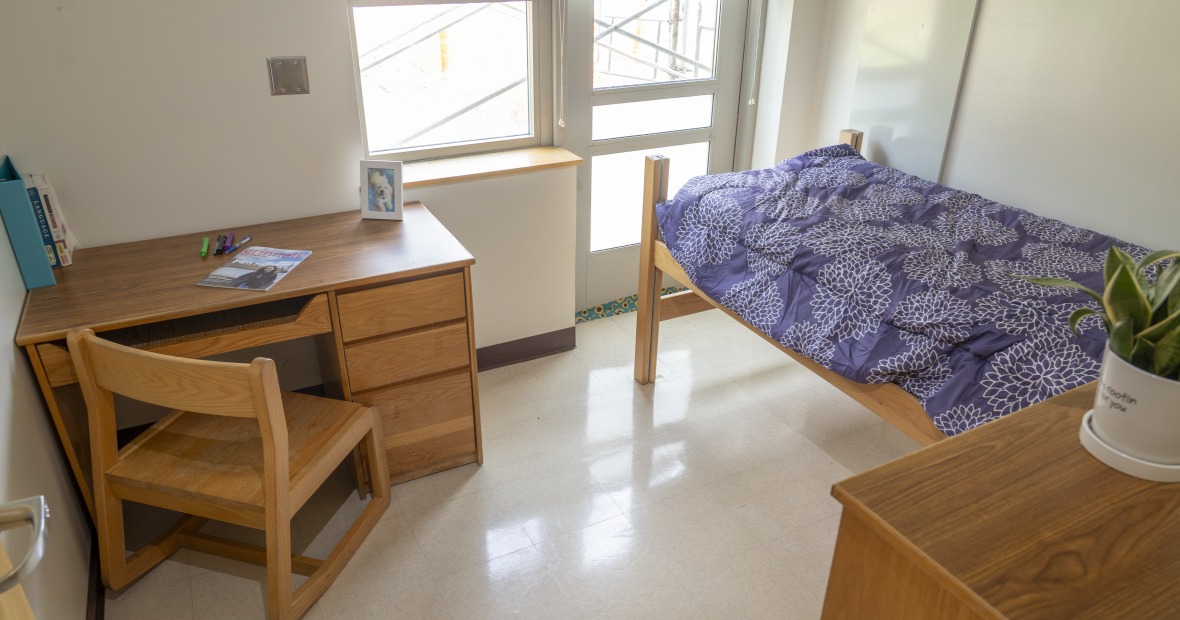 Single bedroom with large windows, a bed, desk, and dresser.