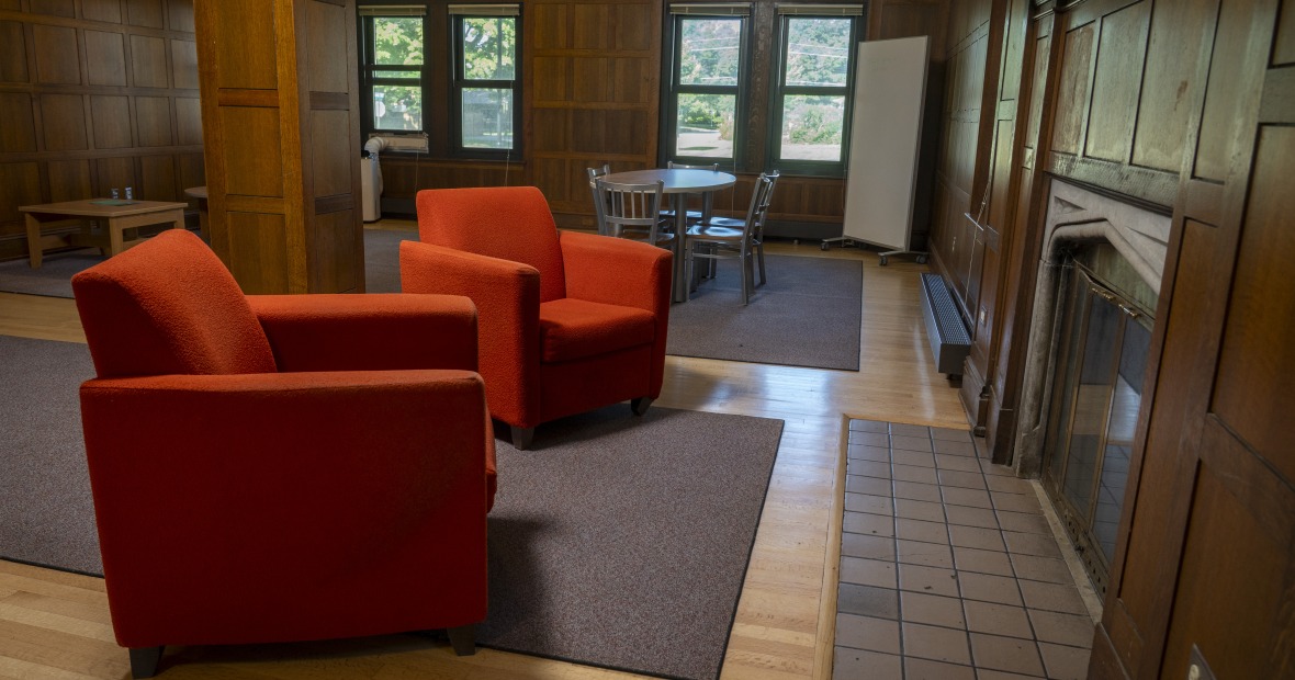 A cozy lounge with tables and plush red chairs in front of a fireplace.