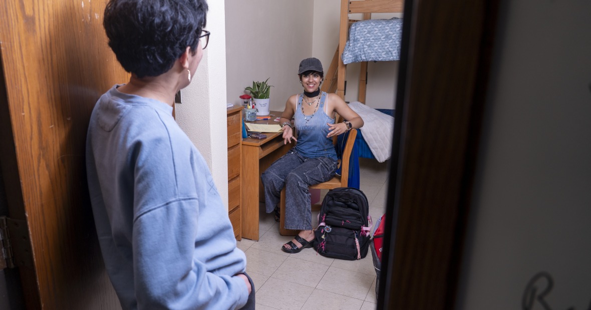 Students chat in a residence hall room. One is at a desk and the other at the door.