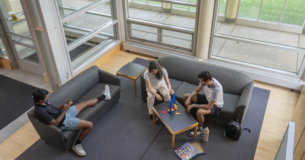 Students sit on couches and play board games at a lounge area surrounded by glass windows..