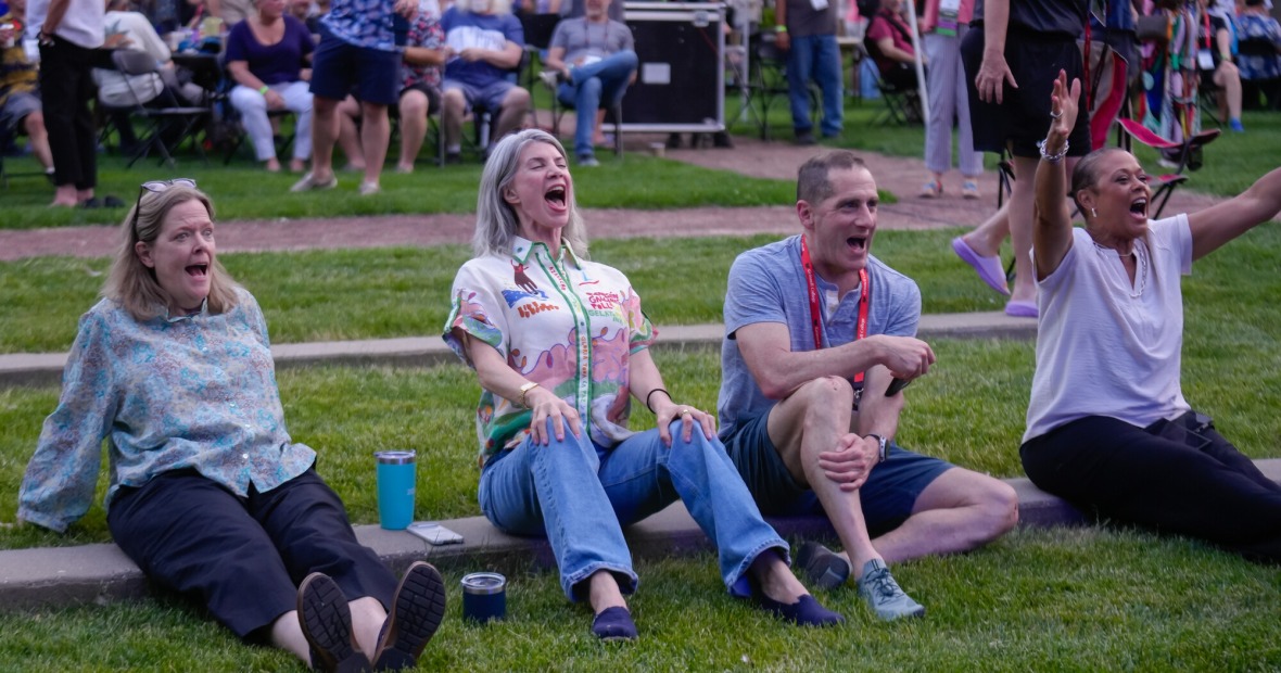 Four alums sitting on the grass cheering for the on stage performance.
