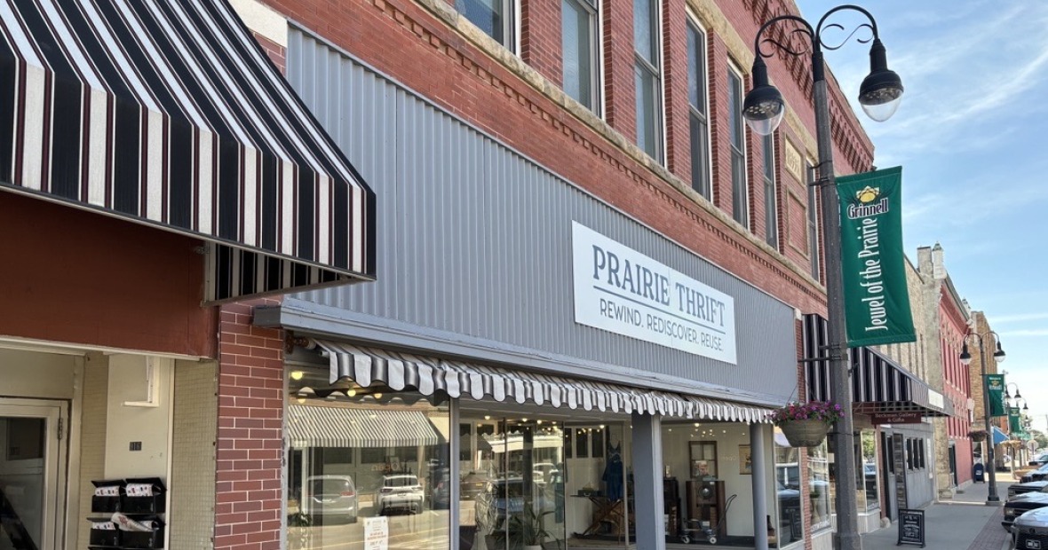 A row of shops with a white sign on a gray storefront reading "Prairie Thrift: Rewind. Rediscover. Reuse."