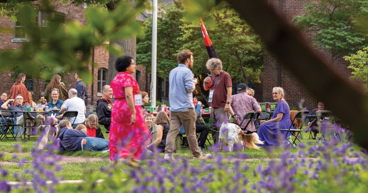 A diverse crowd of Grinnell alumni mingle at reunion against a background of college buildings and colorful flower beds 