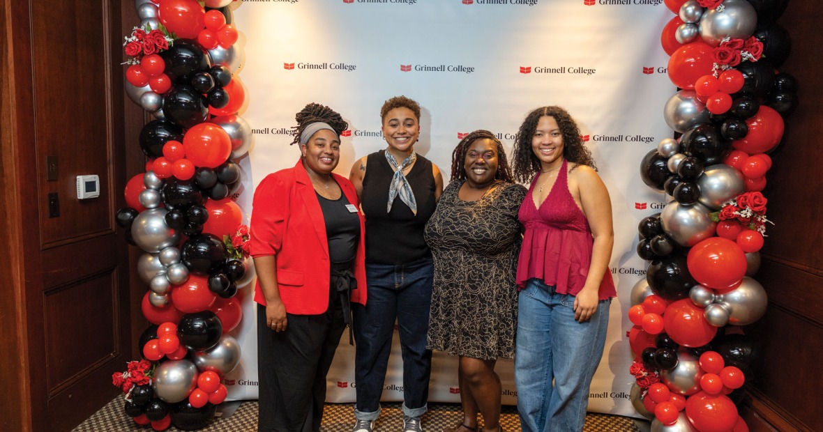 A Group of female black alumni and staff pose in front of a Grinnell College background during Black Alumni Weekend