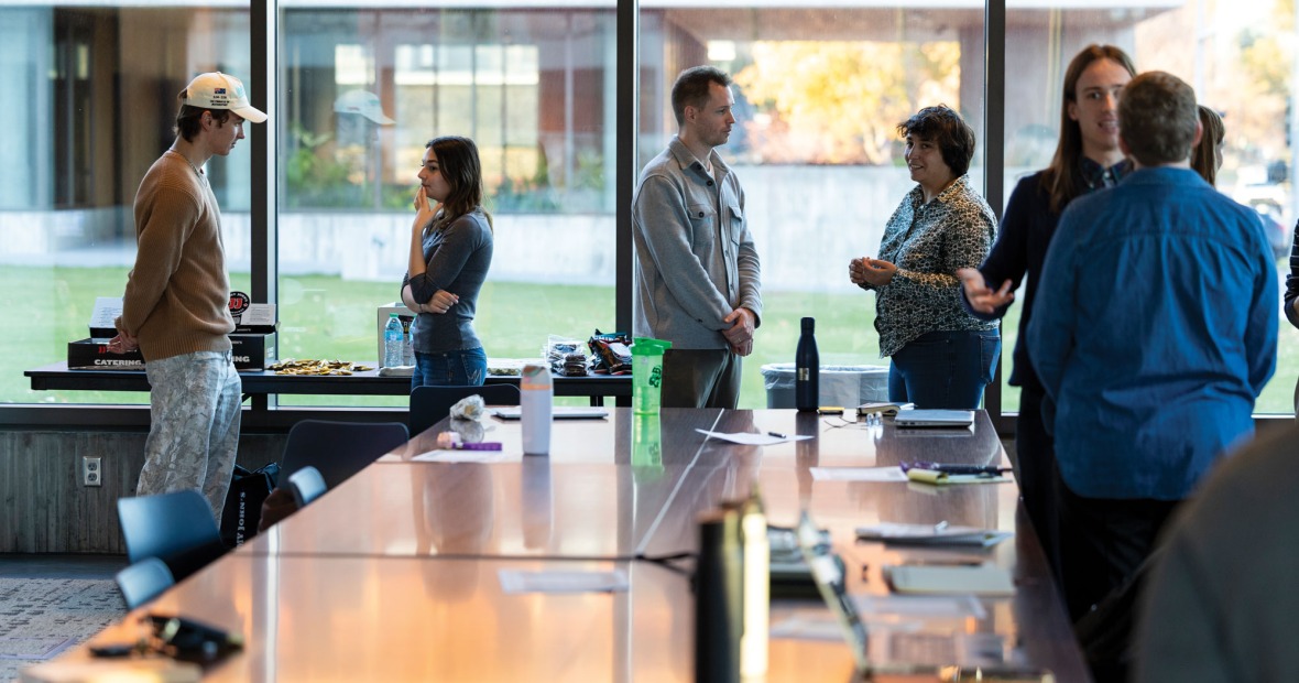 Students and community members are pictured in conversation around a table with a floor to ceiling window in the background 