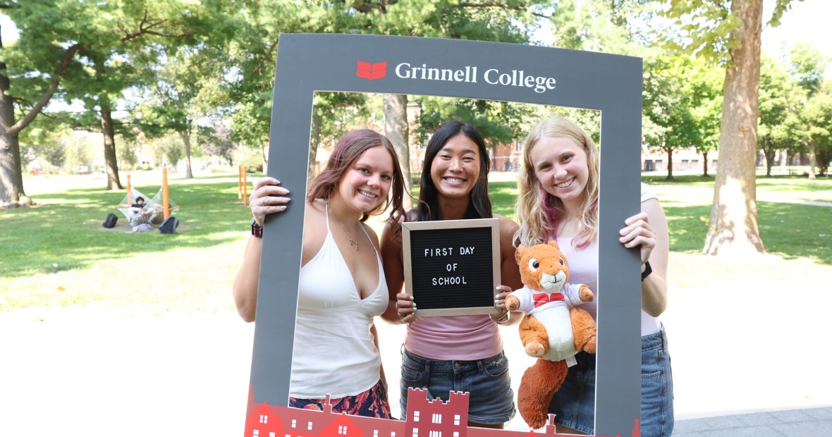 Three students pose for a photo holding a large grey photo frame and a sign that says First Day of School.
